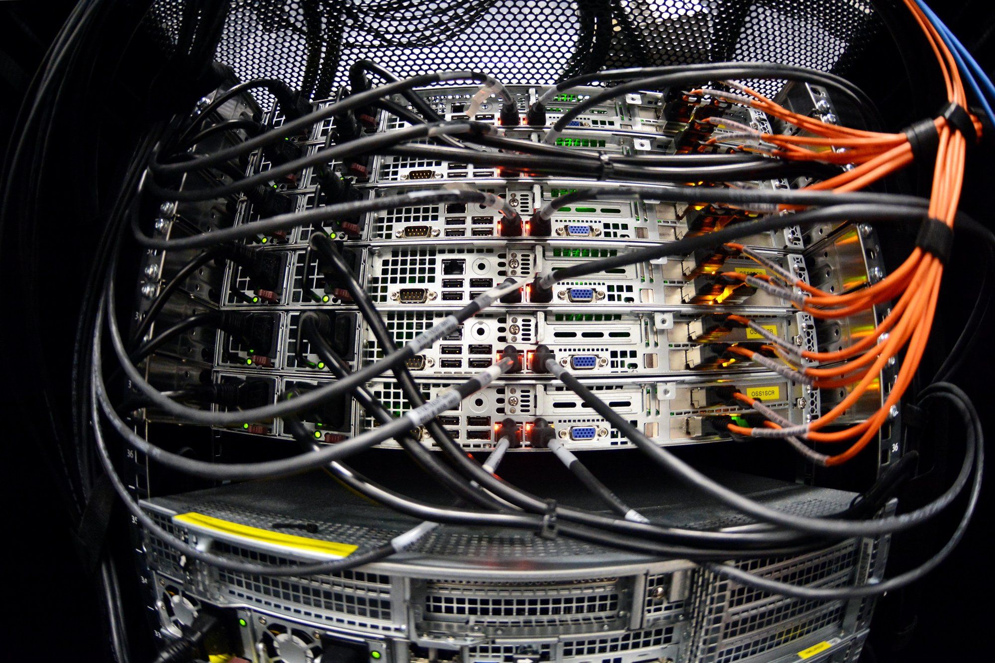 Back of a data-centre server rack, rows of servers laced with black cables and bundles of orange fibre-optic patch cords.